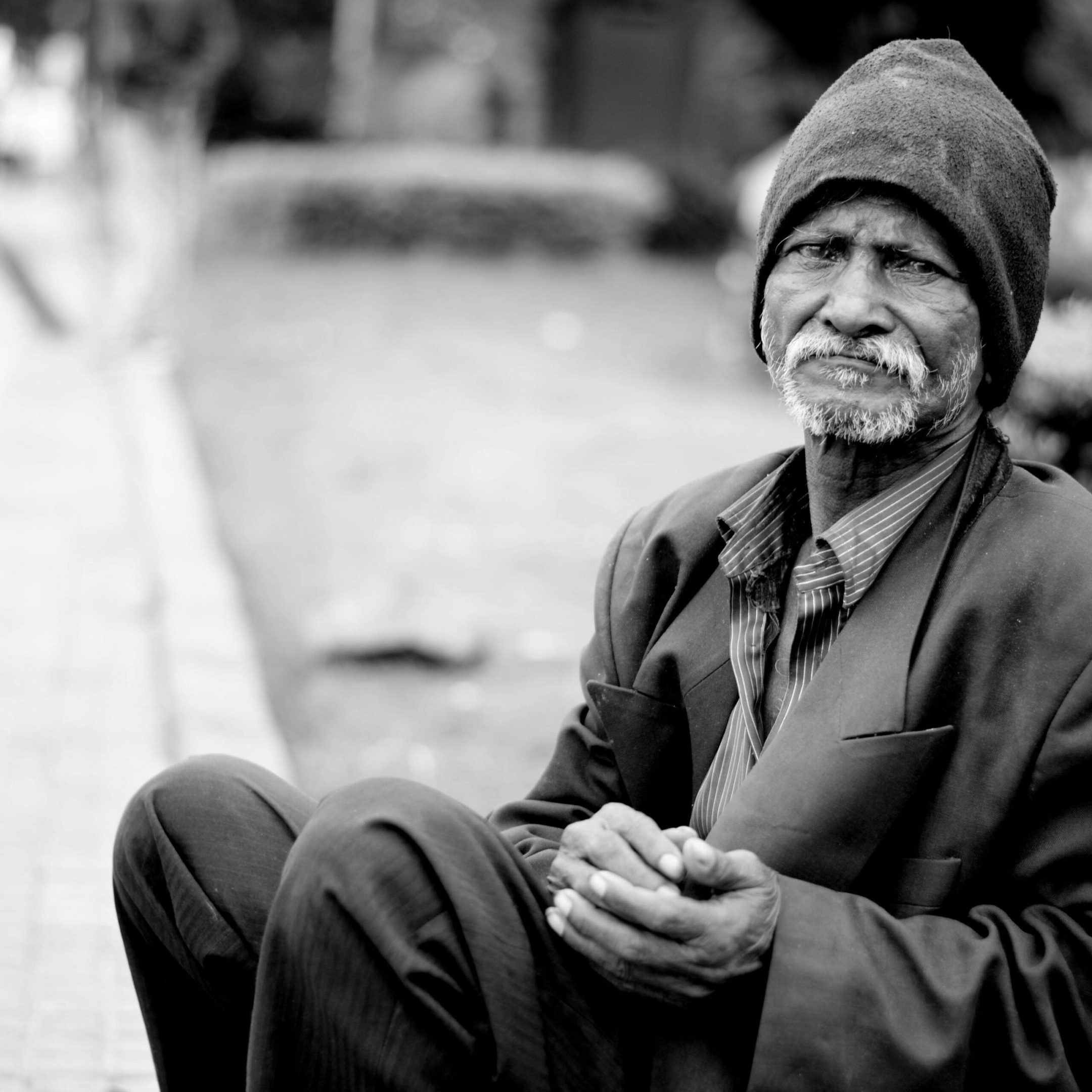 An elderly man in a beanie and coat sitting outdoors, looking thoughtfully at the camera.